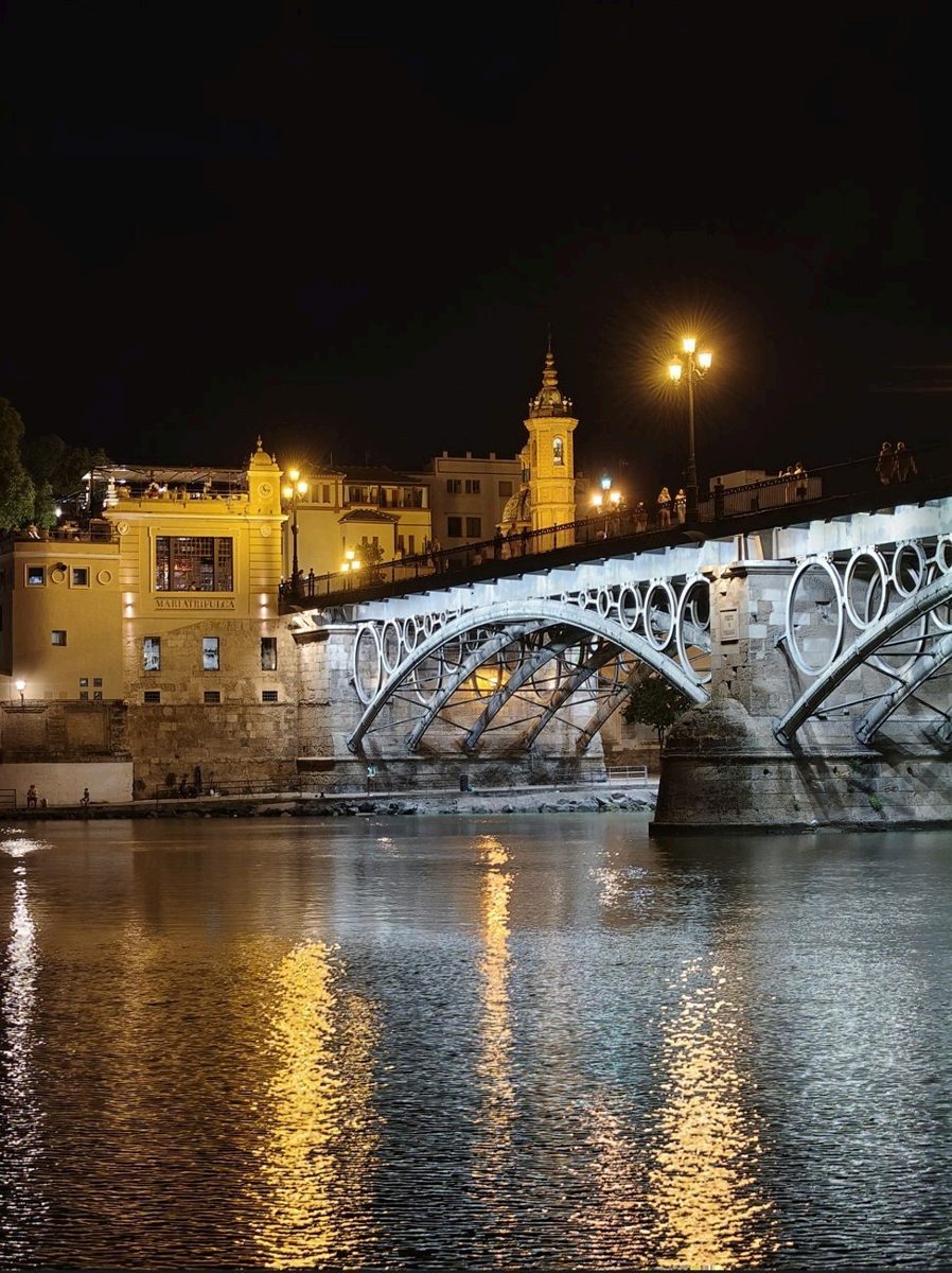Puente de Triana de noche en Sevilla, con los arcos iluminados y el reflejo de las luces en el río
